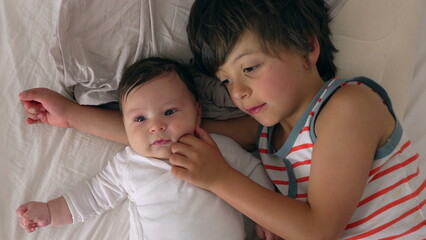 Baby and older sibling lying side by side on bed, sibling gently touching baby’s face, both children bonding in a calm and intimate moment