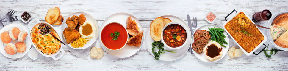 Cozy comfort food table scene. Above view over a white wood banner background. Meatloaf, stew, pot pie, mac and cheese, grilled cheese, soup, fried chicken, shepherds pie and warm sides.