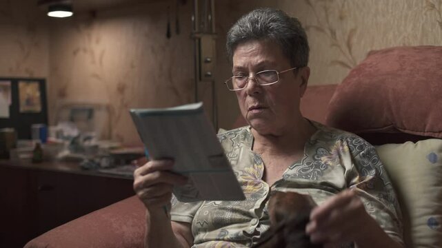 Elderly woman sitting on sofa and solving crossword puzzle, holding dachshund.