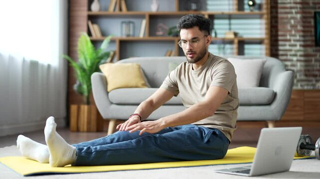 Handsome man practices gymnastics watching online course on laptop sitting on mat on floor in living room at home. Sporty fit male doing stretching exercises listening to instructor via video call