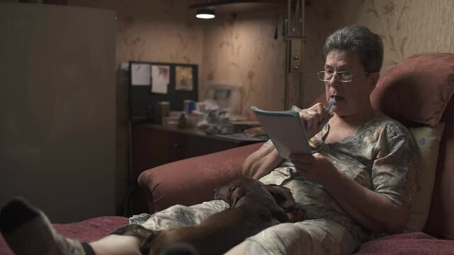 Elderly woman sitting on sofa and solving crossword puzzle, holding dachshund.