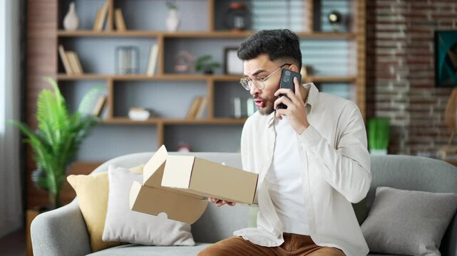 Disappointed young adult man on couch holding phone, expressing frustration about online shopping issue. Open box in front means delivery problem. Unhappy male receiving parcel with wrong product