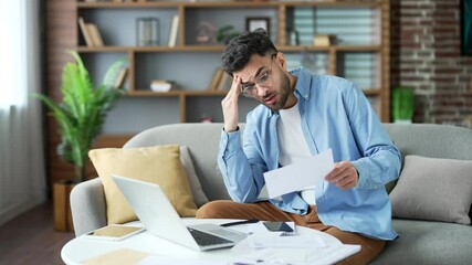 Frustrated young adult man looking at large utility bills sitting on sofa at home office. Upset bearded male holding a paycheck, thinking about problems in the household budget and financial expenses