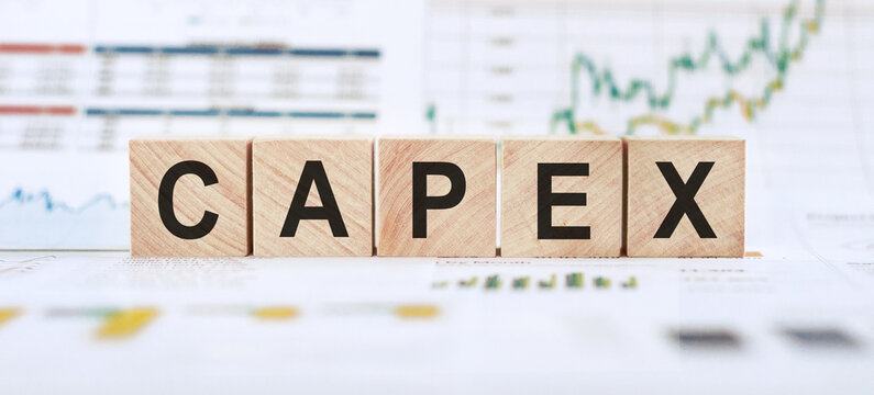 Wooden blocks spelling out CAPEX are arranged on a table with financial charts in the background.