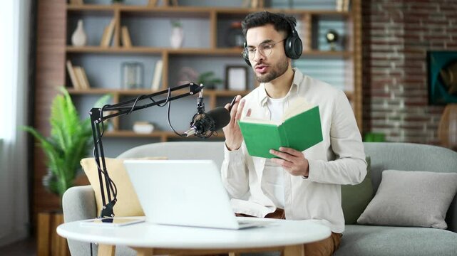 Confident man in wireless headphones recording audiobook using laptop computer and microphone while sitting in home office. Blogger reads a book, conducts a remote conversation on an online video call