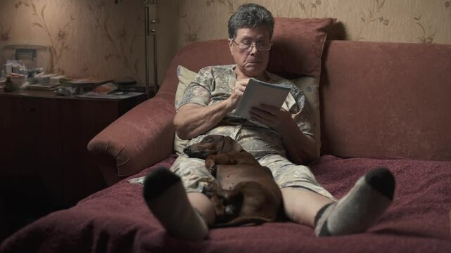 Elderly woman sitting on sofa and solving crossword puzzle, holding dachshund.