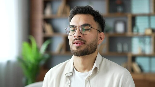 Close up of young adult smiling man relaxing with his eyes closed sitting on sofa in living room at home. Face of calm happy handsome bearded male resting breathing deeply. He feels happiness, relief
