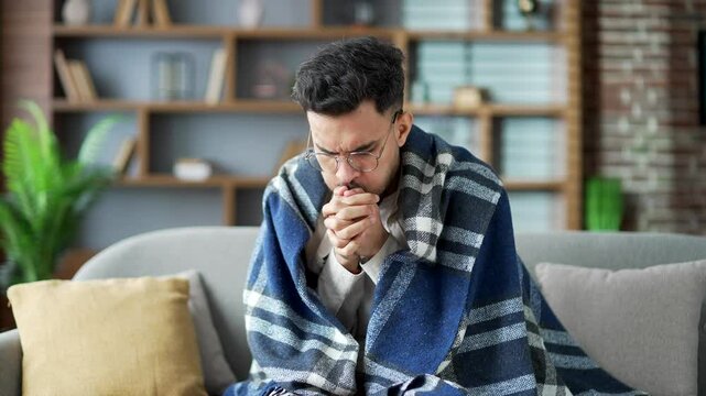 Upset young adult man is frozen covered with blanket sitting on sofa in living room at home. Disappointed male cold indoors and warms his hands by rubbing. Guy feels discomfort due to low temperature