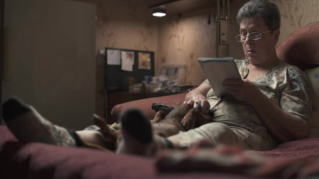Elderly woman sitting on sofa and solving crossword puzzle, holding dachshund.