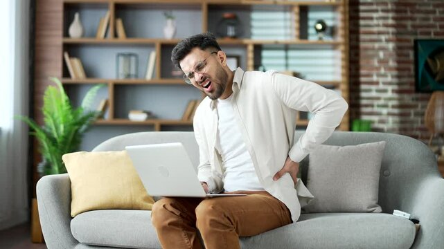 Tired young adult man suffering from back pain sitting on sofa in living room at home. Upset bearded male holds his lower back with his hands, massages painful muscles, has sciatica or a sick spine