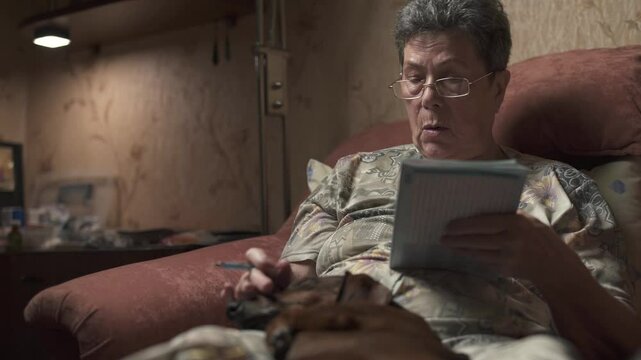 Elderly woman sitting on sofa and solving crossword puzzle, holding dachshund.