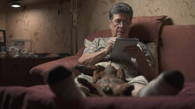 Elderly woman sitting on sofa and solving crossword puzzle, holding dachshund.