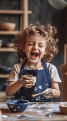 Happy young child with curly hair laughing while playing with clay. The child is wearing an apron and holding a pottery piece, showing joy and creativity. Indoor studio setting with pottery tools and 