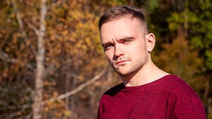 A Young Man in a Maroon Shirt Stands Confidently Against a Backdrop of Autumn Foliage in a Serene Outdoor Setting