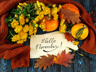 Lettering hello november on kraft paper, pen, pumpkins, orange sweater, autumn leaves, bouquet of yellow chrysanthemum flowers on wooden background © Ivamedia