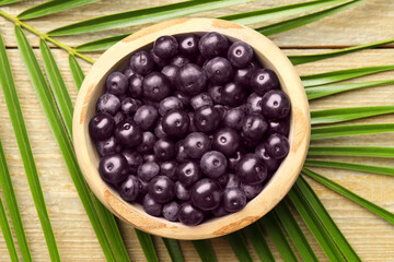 Ripe acai berries in bowl and palm leaves on wooden table, top view
