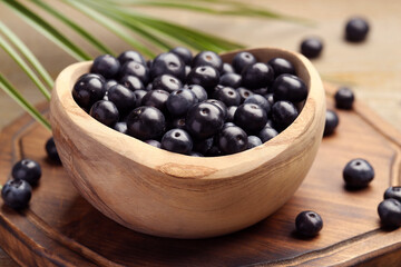 Ripe acai berries in bowl and palm leaves on wooden table, closeup