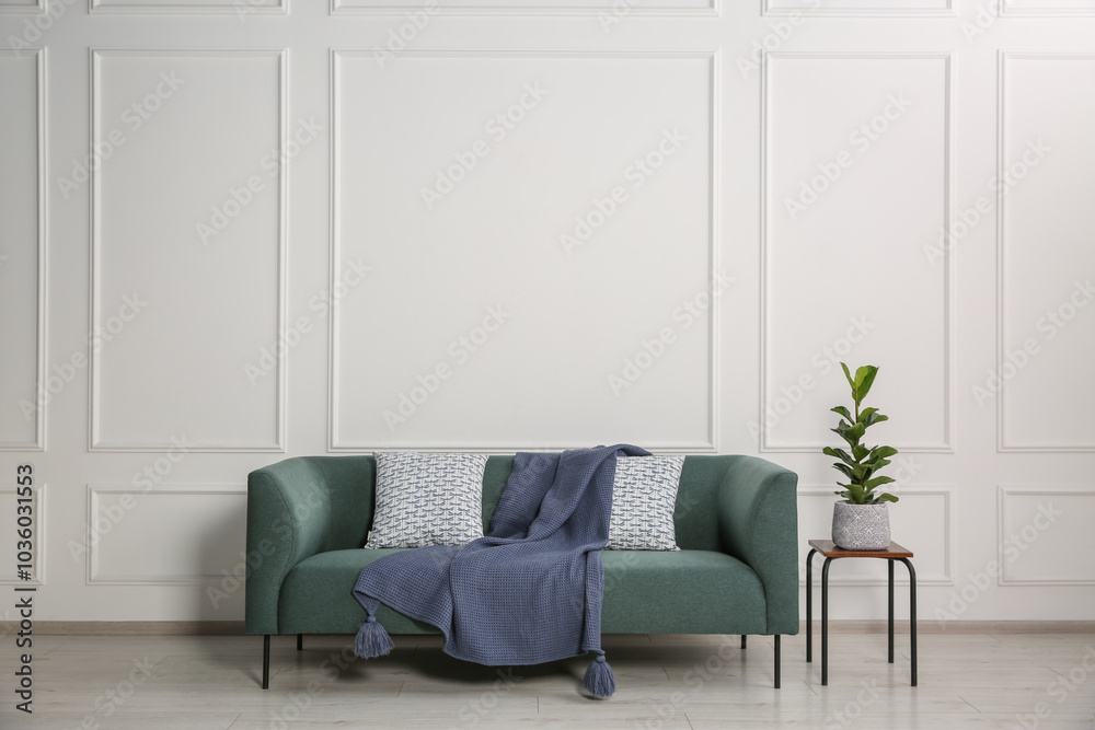 Sticker Stylish sofa with cushions, blanket and plant on side table near white wall