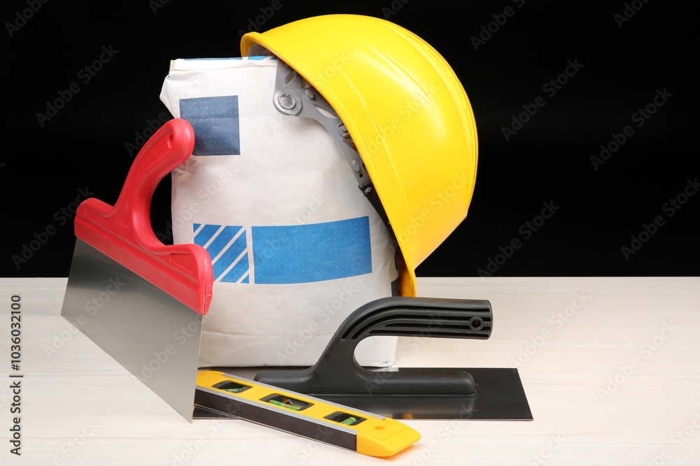 Poster Yellow hard hat, bag of cement powder, putty knives and spirit level on light wooden table against black background. Building material and construction tools