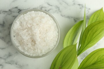 Sea salt in glass bowl and green leaves on white marble table, top view