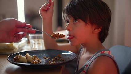 Reluctant young boy being fed by a parent, initially hesitating but then opening his mouth wide to...