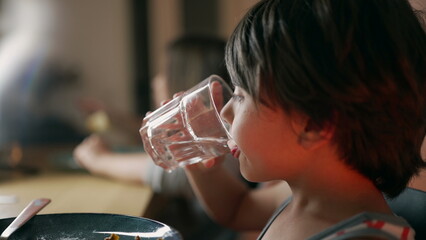 Side profile of a young boy drinking from a glass of water at the dining table, focusing on his...