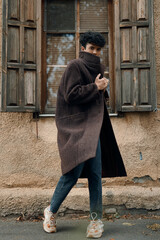 Fashion man in brown coat and jeans walking in front of old building with window in background