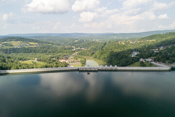 Aerial view of a serene dam surrounded by lush green hills and a calm lake reflecting the blue sky and clouds