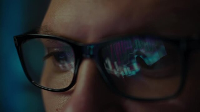 A person with glasses is intently watching a computer screen illuminated by colorful lights. The close-up emphasizes their focused expression and the glow reflecting in their eyes