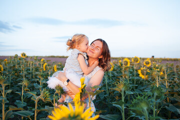 Happy and smiling mother and a little adorable cute blond daughter are hugging and kissing in a sunflower field at sunset