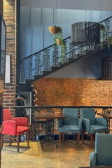 cozy vintage interior of the cafe restaurant with blue walls, wooden floors and brickwork. blue and red chairs are arrenged around tables. natural lighting and lamps. wrought-iron staircase leads up.