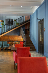 cozy vintage interior of the cafe restaurant with blue walls, wooden floors and brickwork. blue and red chairs are arrenged around tables. natural lighting and lamps. wrought-iron staircase leads up.