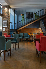 cozy vintage interior of the cafe restaurant with blue walls, wooden floors and brickwork. blue and red chairs are arrenged around tables. natural lighting and lamps. wrought-iron staircase leads up.