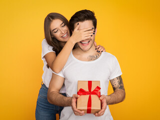 Girl covering her boyfriend eyes and giving him present on yellow background