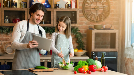 Modern technologies and cooking. Smiling dad and his little daughter reading recipe on digital tablet, preparing food in kitchen together, copy space