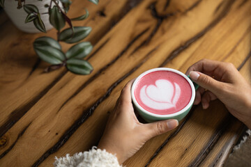 Young woman's hand holds a cup of matcha tea with raspberry or red berries, heart-shaped milk art, cappuccino coffee with red foam and heart, wooden table and decoration with plants, vegan hot drink