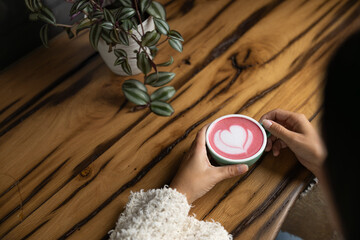 Young woman's hand holds a cup of matcha tea with raspberry or red berries, heart-shaped milk art, cappuccino coffee with red foam and heart, wooden table and decoration with plants, vegan hot drink