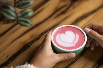 Young woman's hand holds a cup of matcha tea with raspberry or red berries, heart-shaped milk art, cappuccino coffee with red foam and heart, wooden table and decoration with plants, vegan hot drink