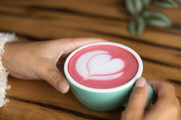 Young woman's hand holds a cup of matcha tea with raspberry or red berries, heart-shaped milk art, cappuccino coffee with red foam and heart, wooden table and decoration with plants, vegan hot drink