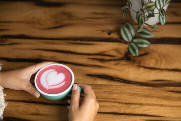 Young woman's hand holds a cup of matcha tea with raspberry or red berries, heart-shaped milk art, cappuccino coffee with red foam and heart, wooden table and decoration with plants, vegan hot drink