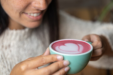 Close-up happy young woman smiling in a cafe holding a cup of matcha tea with raspberry, heart-shaped latte art, cappuccino coffee with red foam and heart, decoration with plants, vegan hot drink
