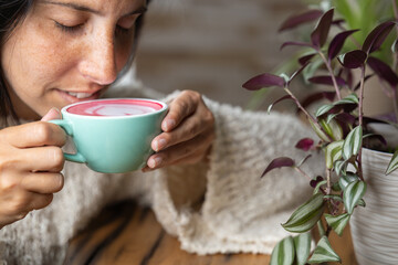 Close-up happy young woman smiling in a cafe holding a cup of matcha tea with raspberry, heart-shaped latte art, cappuccino coffee with red foam and heart, decoration with plants, vegan hot drink