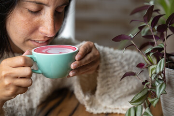 Close-up happy young woman smiling in a cafe holding a cup of matcha tea with raspberry, heart-shaped latte art, cappuccino coffee with red foam and heart, decoration with plants, vegan hot drink