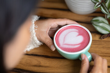 Young woman's hand holds a cup of matcha tea with raspberry or red berries, heart-shaped milk art, cappuccino coffee with red foam and heart, wooden table and decoration with plants, vegan hot drink