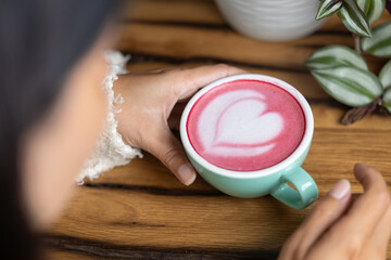 Young woman's hand holds a cup of matcha tea with raspberry or red berries, heart-shaped milk art, cappuccino coffee with red foam and heart, wooden table and decoration with plants, vegan hot drink