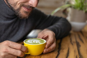 Close-up happy young man smiling in a cafe holding a cup of matcha tea with raspberry, heart-shaped latte art, cappuccino coffee with red foam and heart, decoration with plants, vegan hot drink