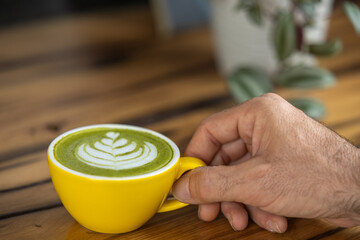 Young man hand holds a cup of matcha tea with raspberry or red berries, heart-shaped milk art, cappuccino coffee with red foam and heart, wooden table and decoration with plants, vegan hot drink