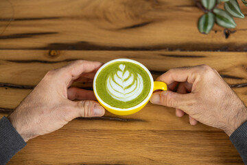Young man hand holds a cup of matcha tea with raspberry or red berries, heart-shaped milk art, cappuccino coffee with red foam and heart, wooden table and decoration with plants, vegan hot drink