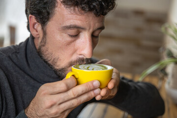 Close-up happy young man smiling in a cafe holding a cup of matcha tea with raspberry, heart-shaped latte art, cappuccino coffee with red foam and heart, decoration with plants, vegan hot drink