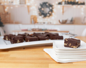 Piece of Homemade Chocolate Pecan Fudge on a White Plate with Tray of Fudge in Background in a Kitchen Decorated for Christmas